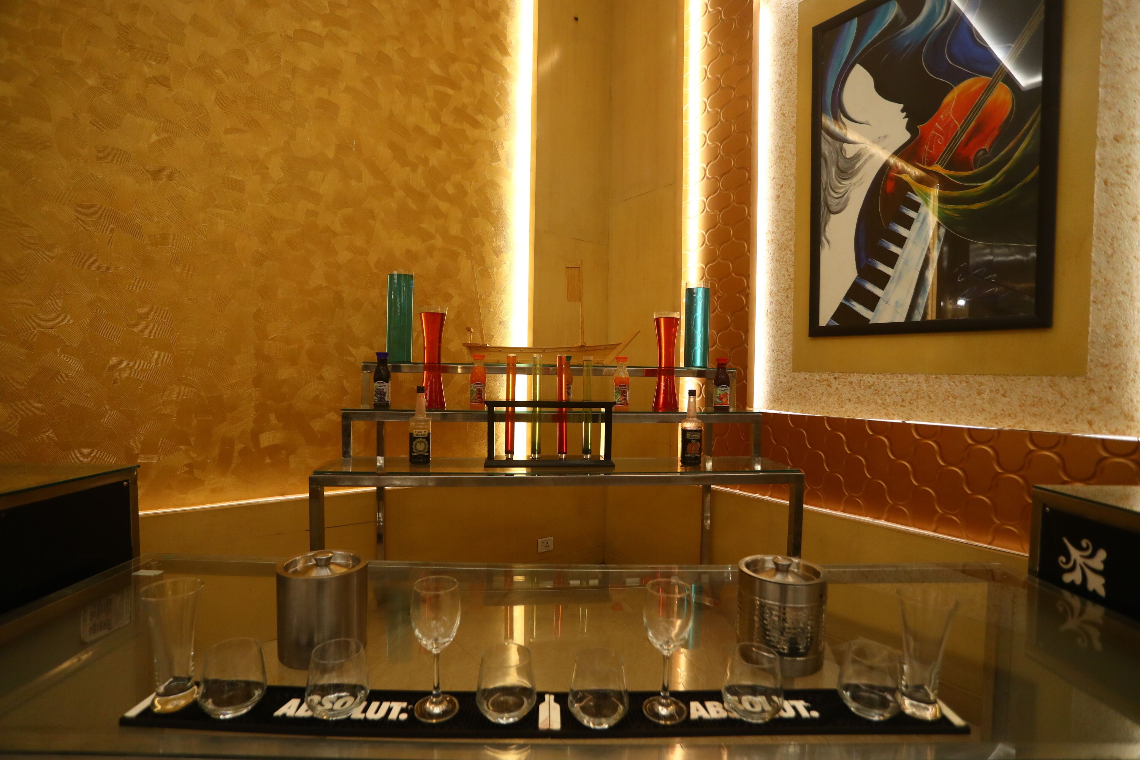 A bar area with a glass counter, glasses, and bottles. There is a painting on the wall and a textured wall behind the bar.