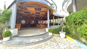 An outdoor seating area with wooden tables and chairs, a curved roof structure, and a paved walkway. - Hotel Kanha's Palm Springs (Bhopal)