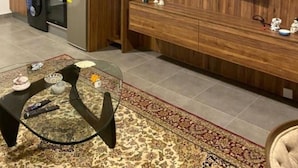Cozinha moderna com armários de madeira, forno embutido e micro-ondas. Há uma televisão de tela plana na parede, uma mesa pequena com objetos decorativos e um tapete estampado no chão.