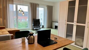 A modern living room with a wooden table, a laptop, a coffee pot, and a mug. There is a green chair, a sofa, and a cabinet in the background.