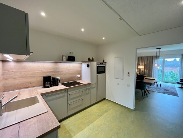 A modern kitchen with a built-in oven, a coffee maker, and a sink. There is a dining area with a table and chairs visible through an open door.