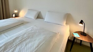 Un lit bien fait, avec du linge de lit blanc, deux lampes de chevet et une petite table sur laquelle repose un livre.