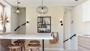 Een moderne keuken met een wit aanrecht, twee houten krukken, hanglampen, een boekenkast en een handdoekhouder.