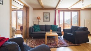 A living room with a sofa, a coffee table, and a view of mountains through large windows. - Living room with eating area, kitchen with dishwasher, Nespresso machine and microwave, one bedroom (Verbier)