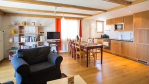 A modern living room with a sofa, dining table, and chairs, a kitchen with wooden cabinets, and a TV. - Living room with eating area, kitchen with dishwasher, Nespresso machine and microwave, one bedroom (Verbier)