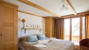A bedroom with a large bed, wooden beams, a balcony with curtains, and a hanging lamp. - Living room with eating area, kitchen with dishwasher, Nespresso machine and microwave, one bedroom (Verbier)