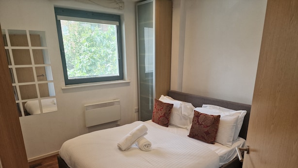 A neatly made bed with white linens and two decorative pillows, a window with blinds, a built-in shelf, and a radiator. - Immaculate 1-bed Apartment in London (London)