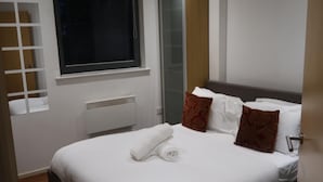 A neatly made bed with a rolled towel, a window with blinds, a radiator, and a door. - Immaculate 1-bed Apartment in London (London)