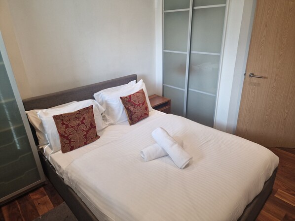 A neatly made bed with white linens and two decorative pillows, a wooden nightstand, and a wooden wardrobe with frosted glass doors. - Immaculate 1-bed Apartment in London (London)