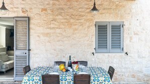 An outdoor dining area with a stone wall, a table set with a tablecloth, chairs, and a bottle of wine.