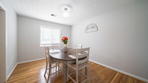 Un coin repas comprenant une table ronde, quatre chaises, un vase avec des fleurs, une fenêtre avec des stores et un luminaire fixé au mur.