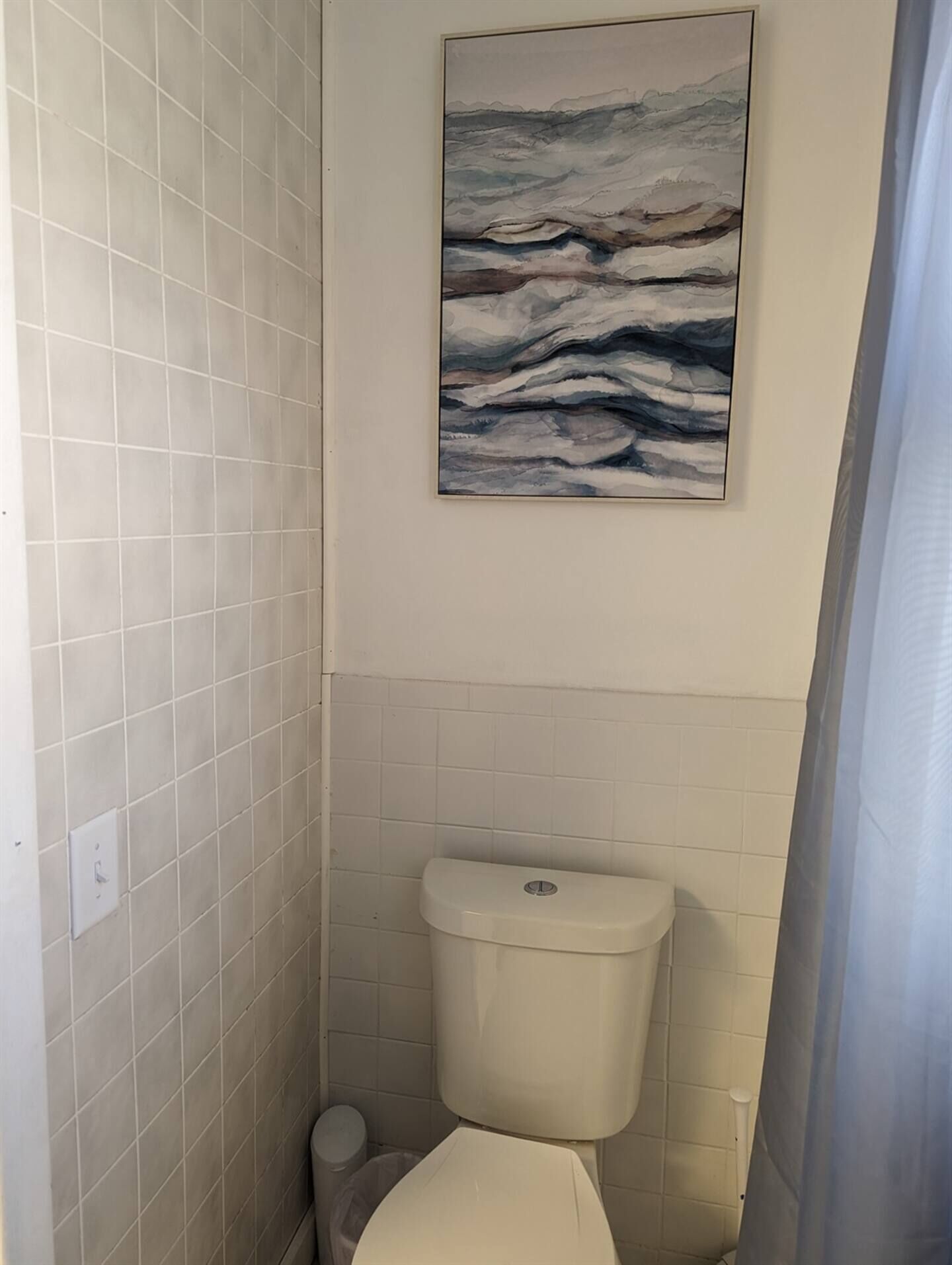 A bathroom with white tiled walls, a toilet, and a wall-mounted abstract painting.