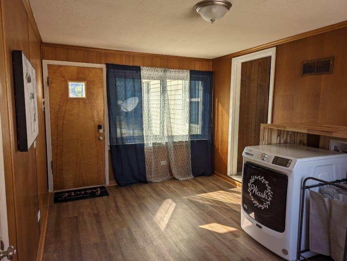 A compact room with wooden walls, a door, a window with curtains, and a washing machine.