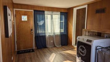 A compact room with wooden walls, a door, a window with curtains, and a washing machine.