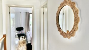 Um corredor com um espelho emoldurado em madeira na parede, uma cama com cobertura estampada de flores e uma cadeira com almofada preta.