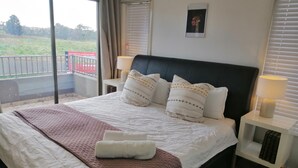 Cama bem arrumada com cobertura acolchoada, duas luminárias de cabeceira e vista para uma varanda com guarda-corpo.