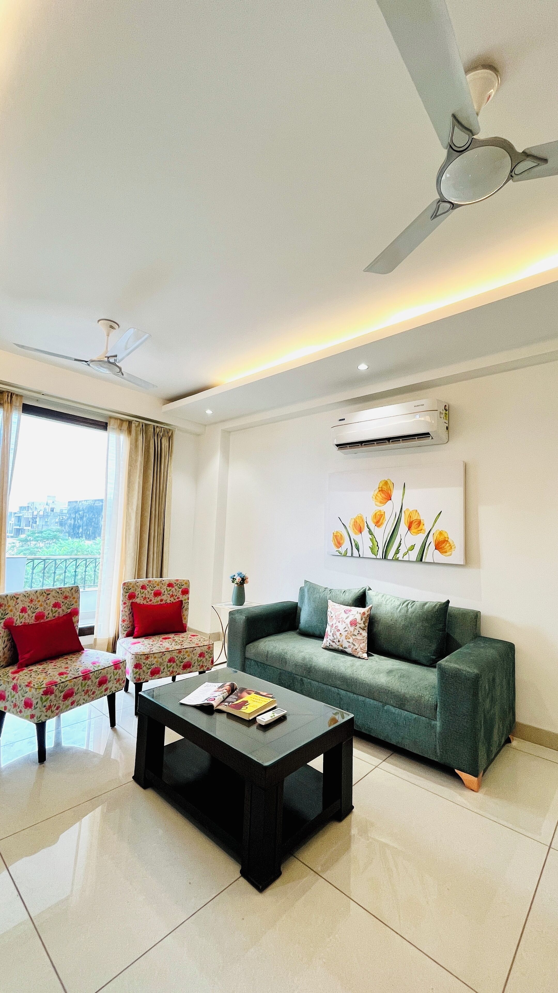 A modern living room with a green sofa, floral armchairs, and a coffee table. There is a ceiling fan, an air conditioning unit, and a window with curtains.