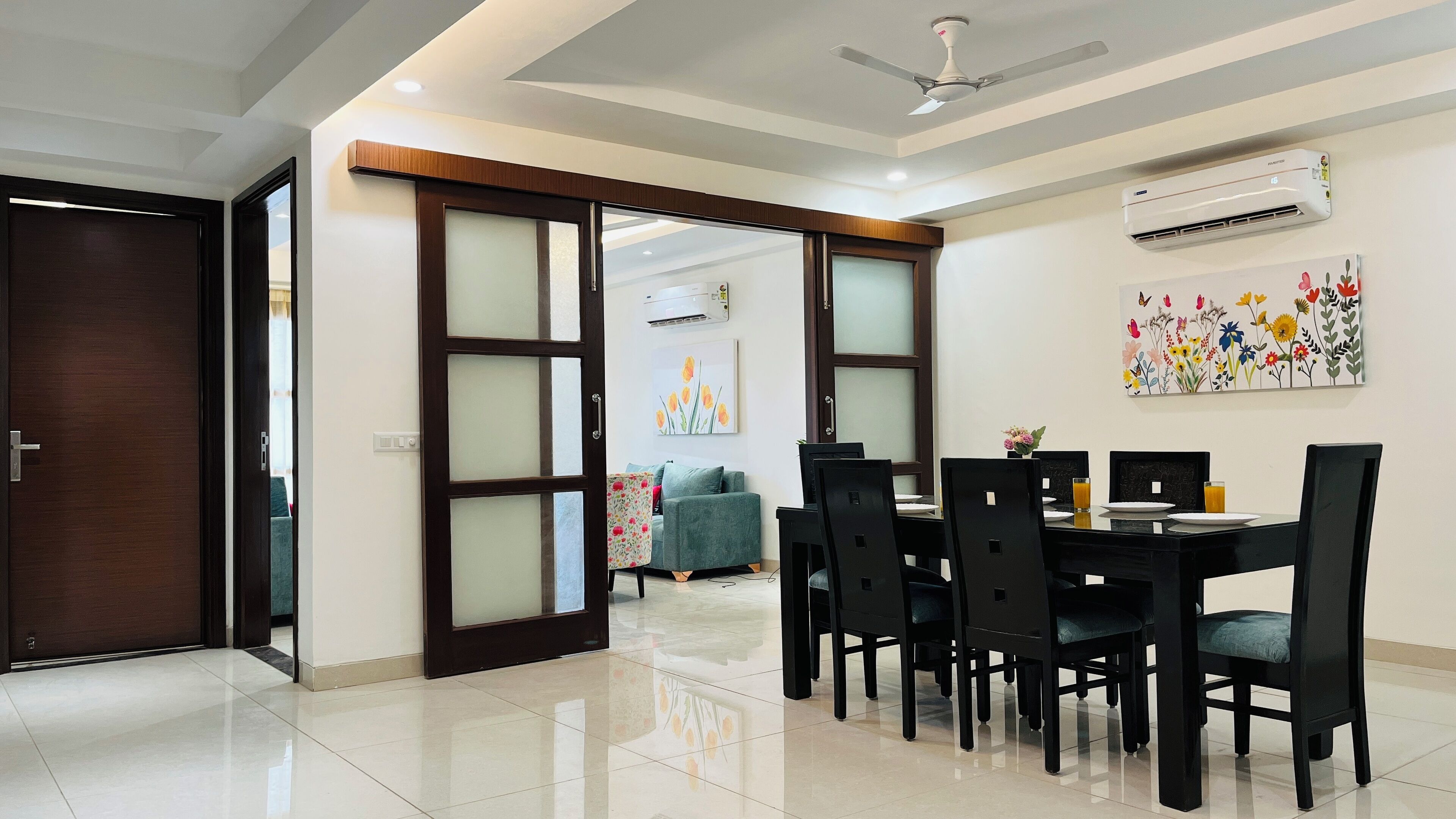 A dining area with a black table and chairs, an air conditioning unit, and a floral painting on the wall.