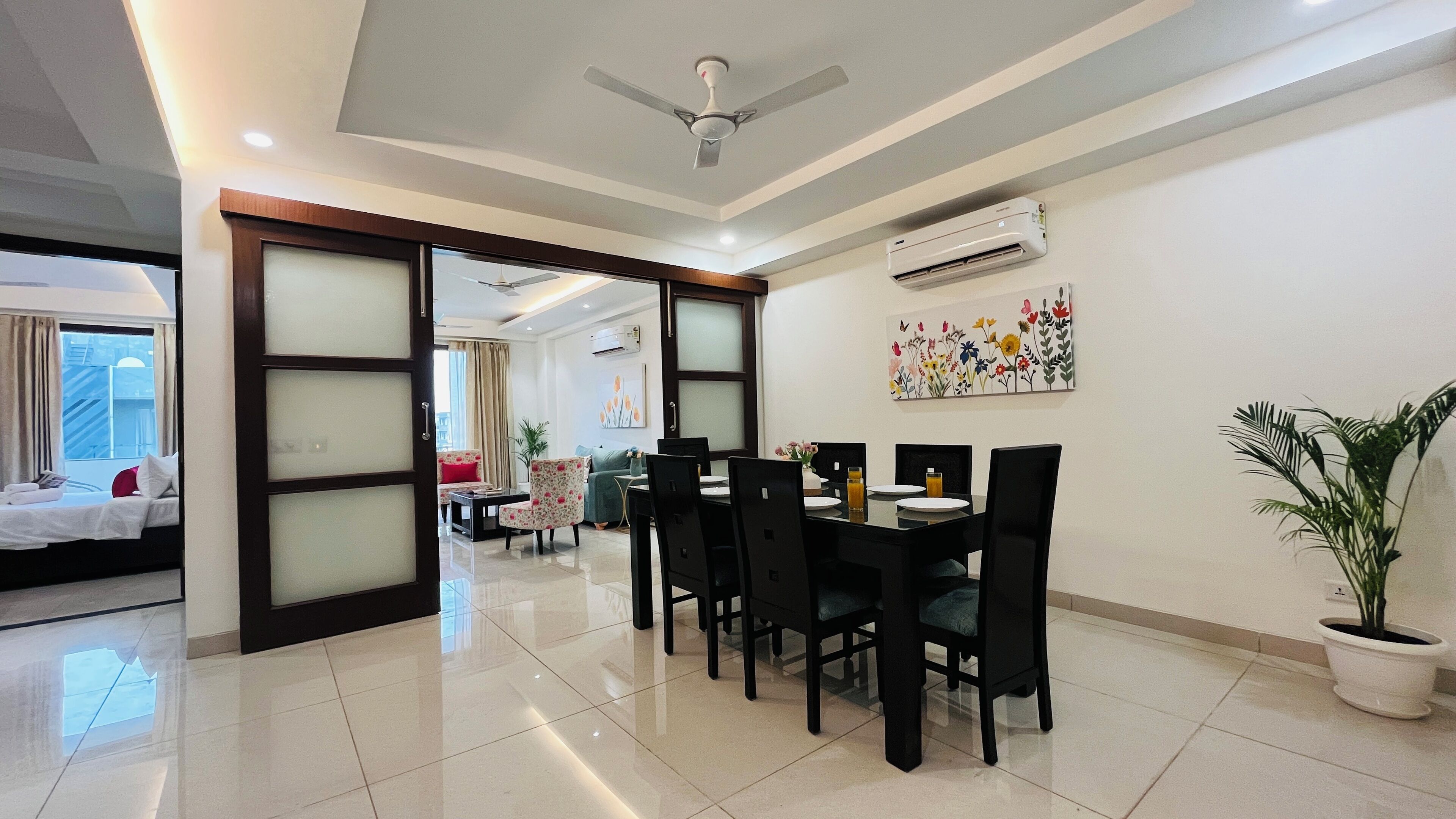 A hotel room with a dining area, a ceiling fan, an air conditioning unit, and a potted plant.