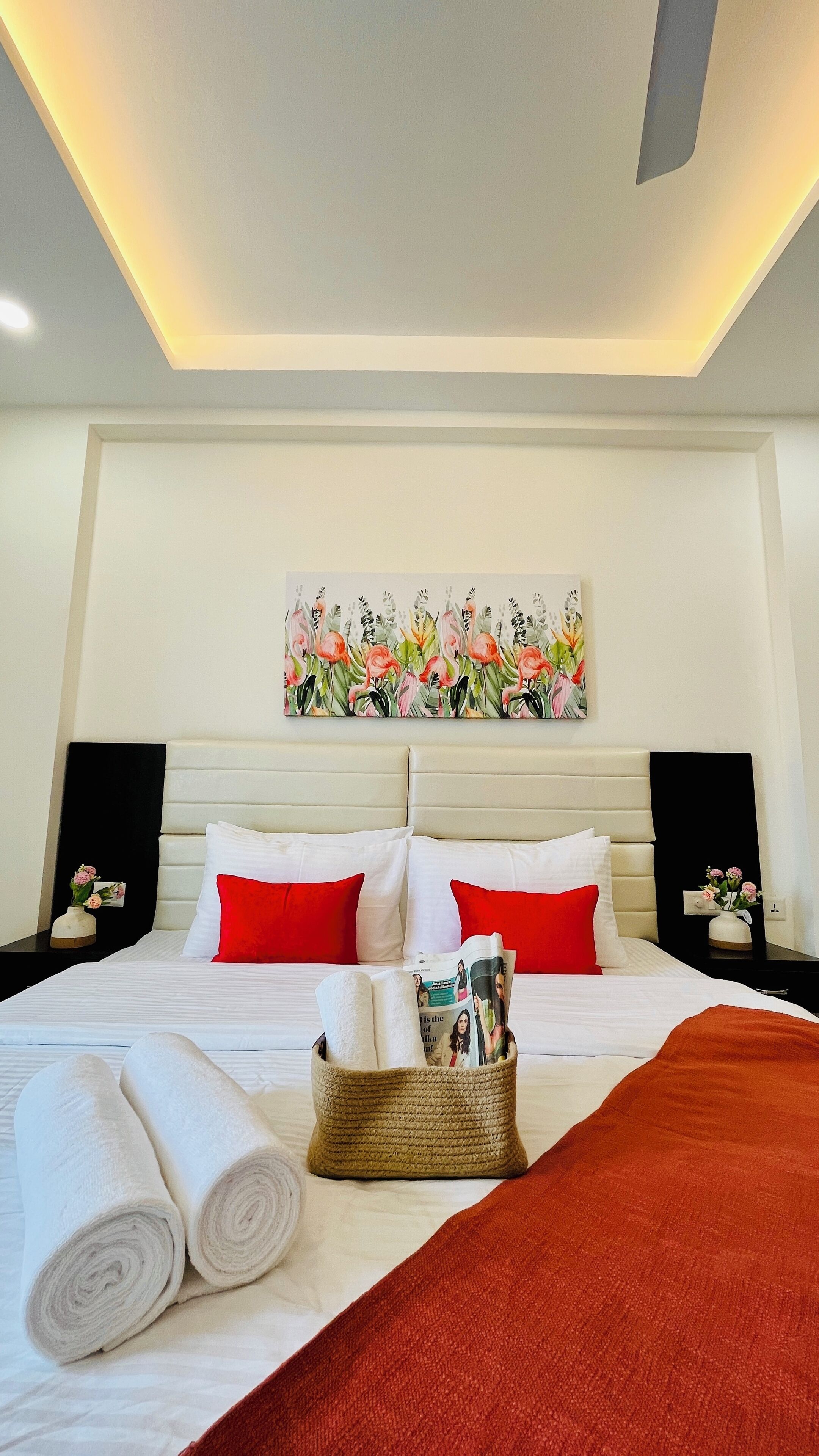 A hotel room with a bed, pillows, a headboard, a nightstand, a vase with flowers, a wall art, and a ceiling light.