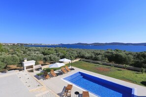 A swimming pool with lounge chairs and umbrellas, surrounded by greenery and a view of the sea. - Villa Capan Zadarvillas (Sveti Filip i Jakov)