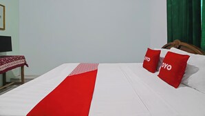 Quarto de hotel com uma cama, dois travesseiros vermelhos, um cobre-leito vermelho e branco, uma mesa de cabeceira de madeira, uma televisão e uma cortina verde.