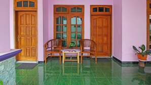 Uma área de recepção com balcão de madeira, duas cadeiras de madeira, uma pequena mesa com uma planta e uma grande porta de madeira com painéis de vidro.