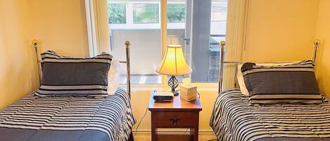 Une chambre avec deux lits, une table de chevet avec une lampe et une fenêtre donnant sur des immeubles.