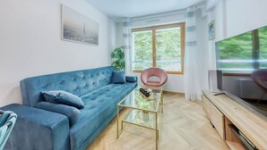 Ein modernes Wohnzimmer mit einem blauen Sofa, einem Glastisch, einem rosa Sessel, einem Fernseher und einem großen Fenster mit Blick auf Grünflächen.