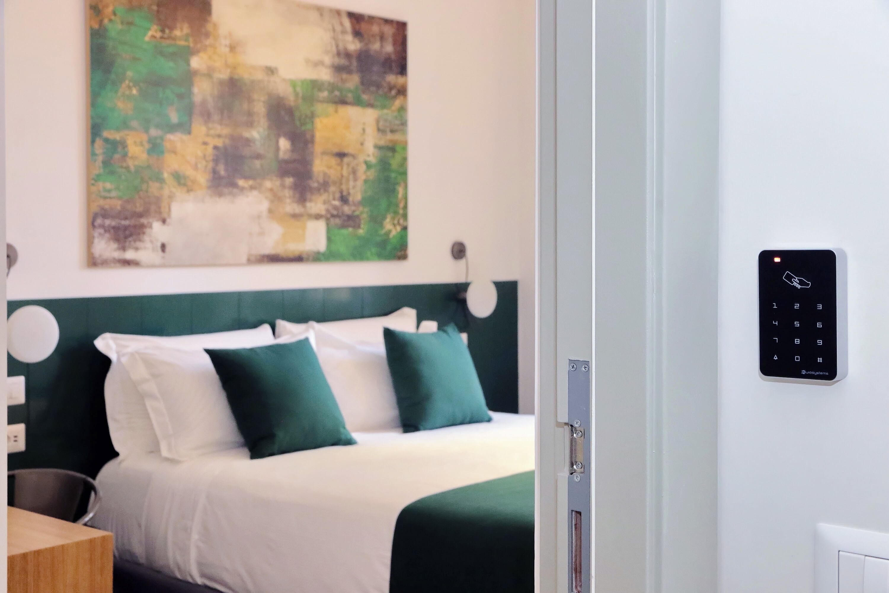 A hotel room with a bed, green pillows, a wall-mounted control panel, and a painting.