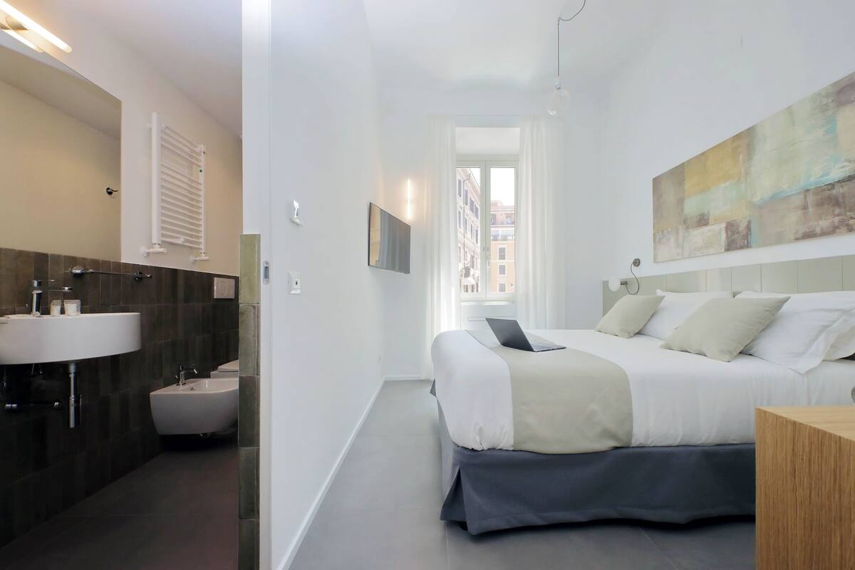 A modern hotel room with a bed, a bedside table, a large window, a bathroom with a sink and bidet, and a city view.