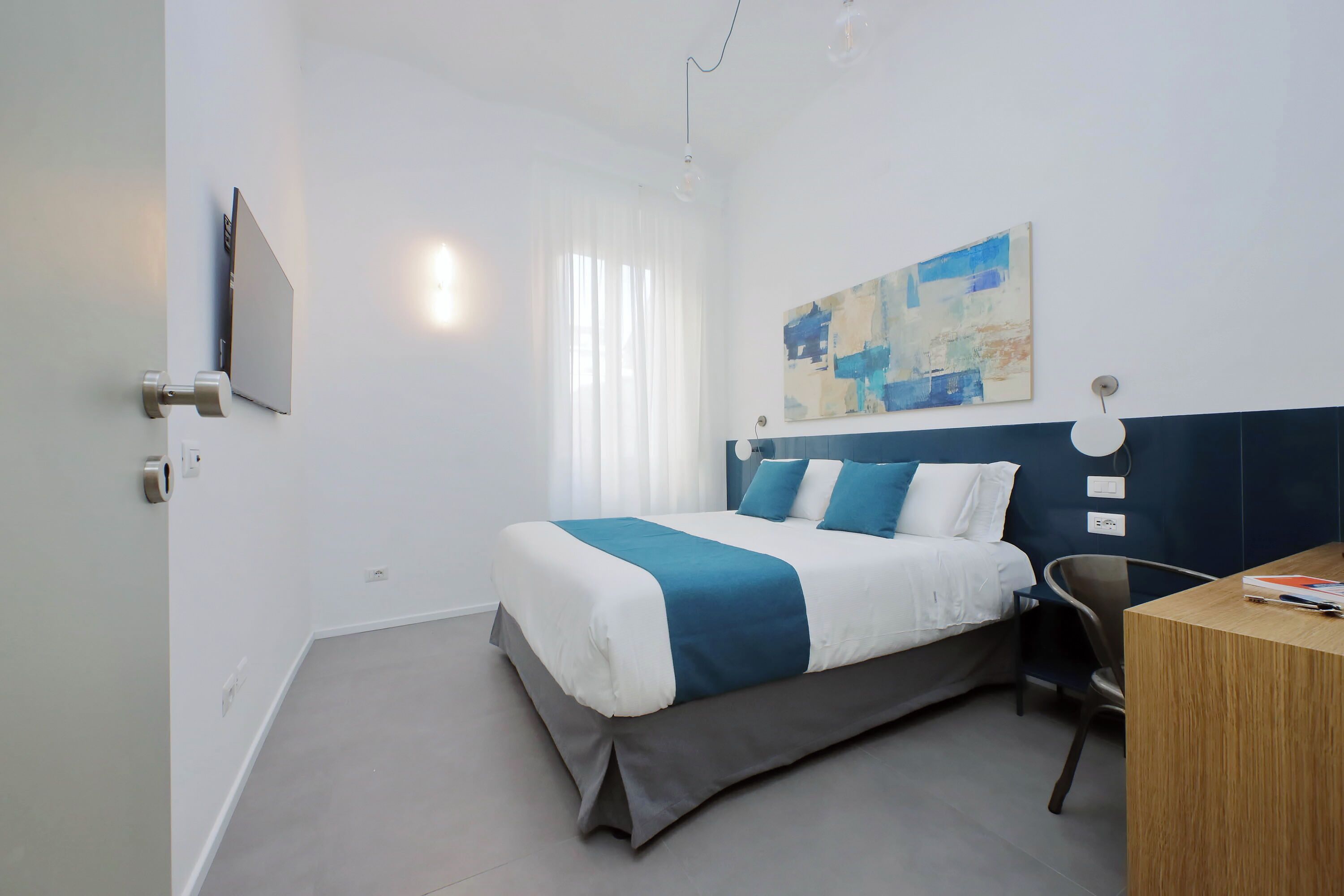 A modern hotel room with a bed, a desk, and a painting on the wall.