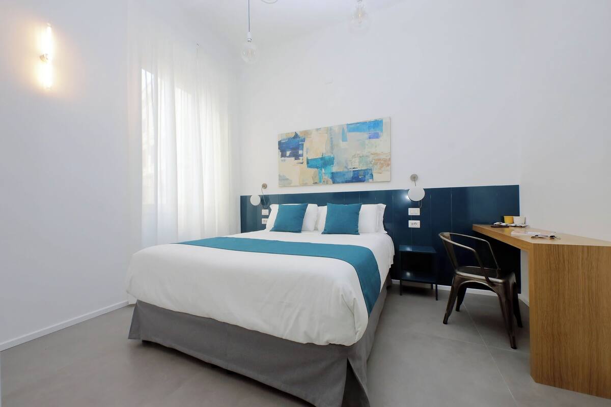 A hotel room with a bed, a desk, a chair, and a painting on the wall.