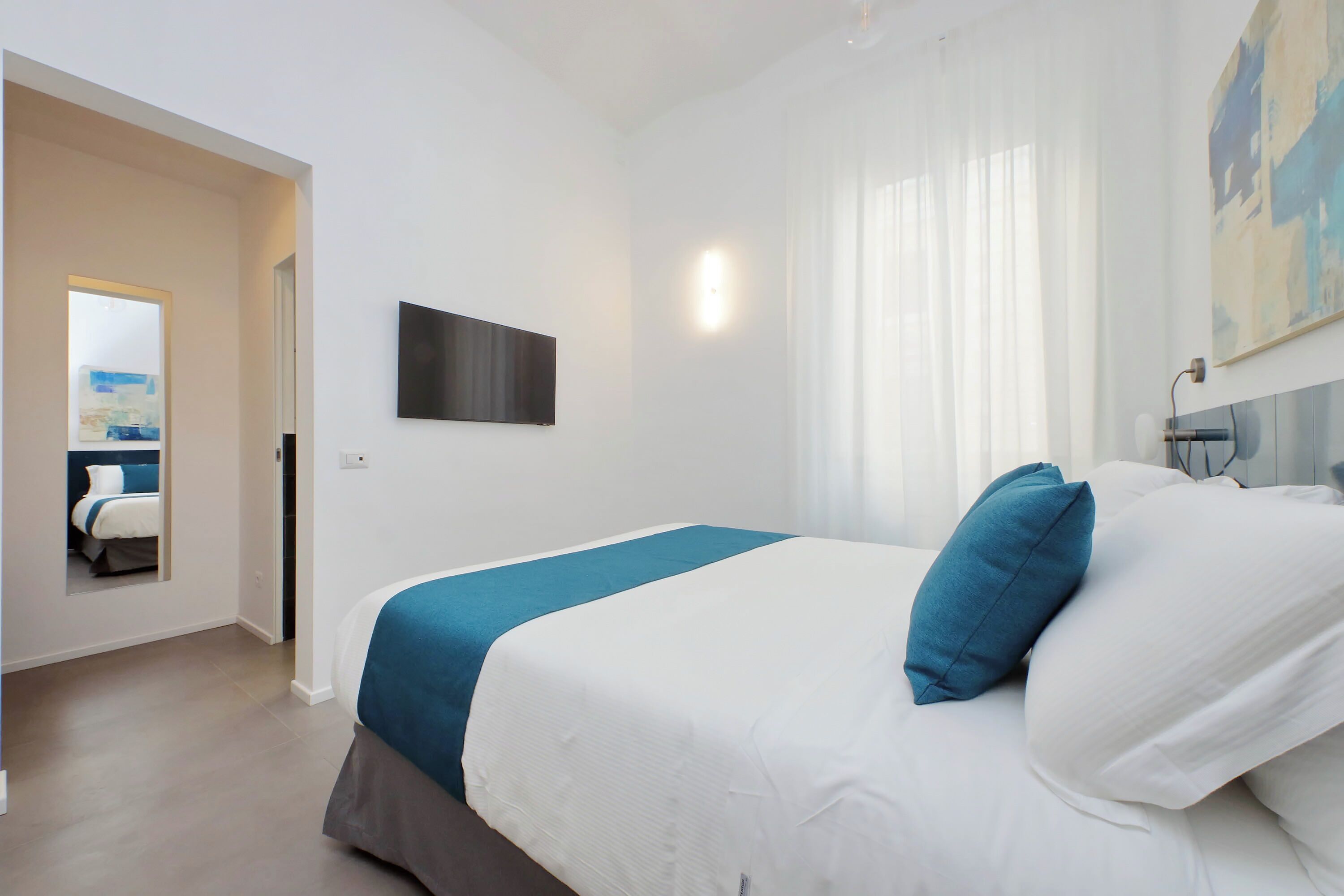 A modern hotel room with a large bed, a flat-screen TV mounted on the wall, and a view of the adjacent room.
