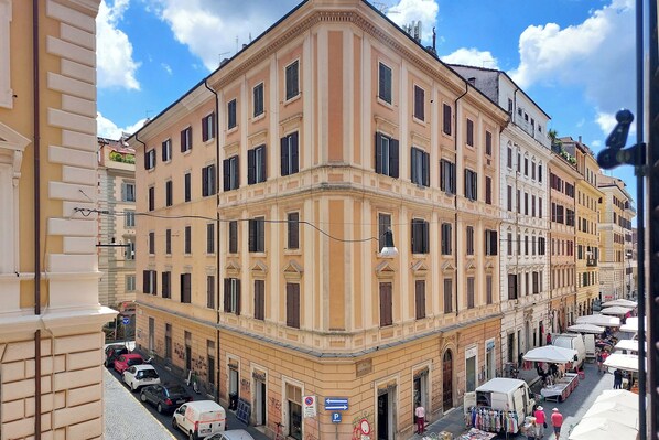 Un edificio de esquina con varios pisos, amplias ventanas y una calle frente a él con coches estacionados y puestos de mercado.