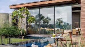 A modern outdoor pool area with a glass wall, lounge chairs, and a dining table. - MYSA Zinc Journey by The Fern (A Glade One Golf Resort) Nani Devati, Gujarat  (Sanand)