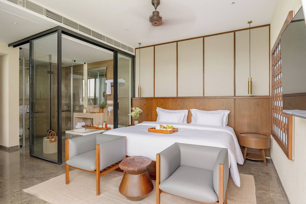 Ein modernes Hotelzimmer mit einem großen Bett, zwei Sesseln, einem kleinen Holztisch und einer Glastür zum Badezimmer.