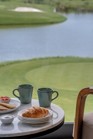 Una mesa con un croissant, un plato de tortitas y dos tazas, con vista a un campo de golf y una masa de agua.