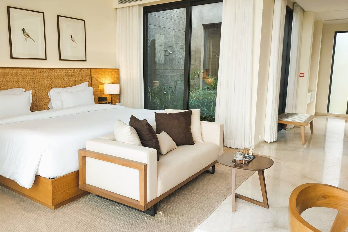 Ein Hotelzimmer mit einem großen Bett, einer Couch, einem kleinen Tisch und einem Sessel. An der Wand befinden sich Bilder in Rahmen, ein Fenster mit Vorhängen und ein Blick ins Grüne.