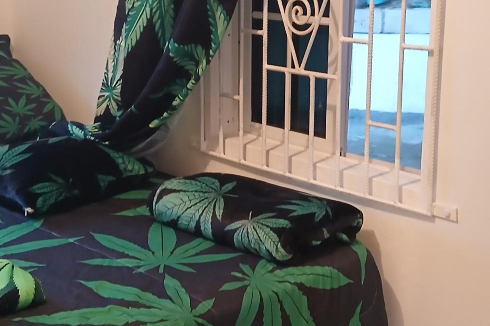 Una cama con una colcha oscura con diseños de hojas verdes, una celosía de metal blanca y vista al mar a través de la ventana.