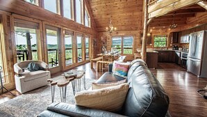 A spacious wooden cabin with large windows, a cozy living area with a leather couch, a dining table, and a kitchen with modern appliances. - 180 Mtn Views! Pool&Pond Access! Sleep 16! Hot tub! Firepit! Prime Location! (Sevierville)