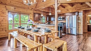 A wooden cabin interior with a dining table set for six, a kitchen with modern appliances, and large windows offering a view of greenery. - 180 Mtn Views! Pool&Pond Access! Sleep 16! Hot tub! Firepit! Prime Location! (Sevierville)
