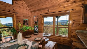 A wooden cabin with a dining table set for four, a large window with a view of mountains, and a wooden ceiling. - 180 Mtn Views! Pool&Pond Access! Sleep 16! Hot tub! Firepit! Prime Location! (Sevierville)