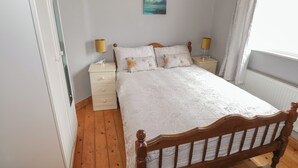 A bedroom with a wooden bed, two bedside tables with lamps, a nightstand, a window with curtains, and a painting on the wall. - 7 Aikenhead Terrace (Foxford)