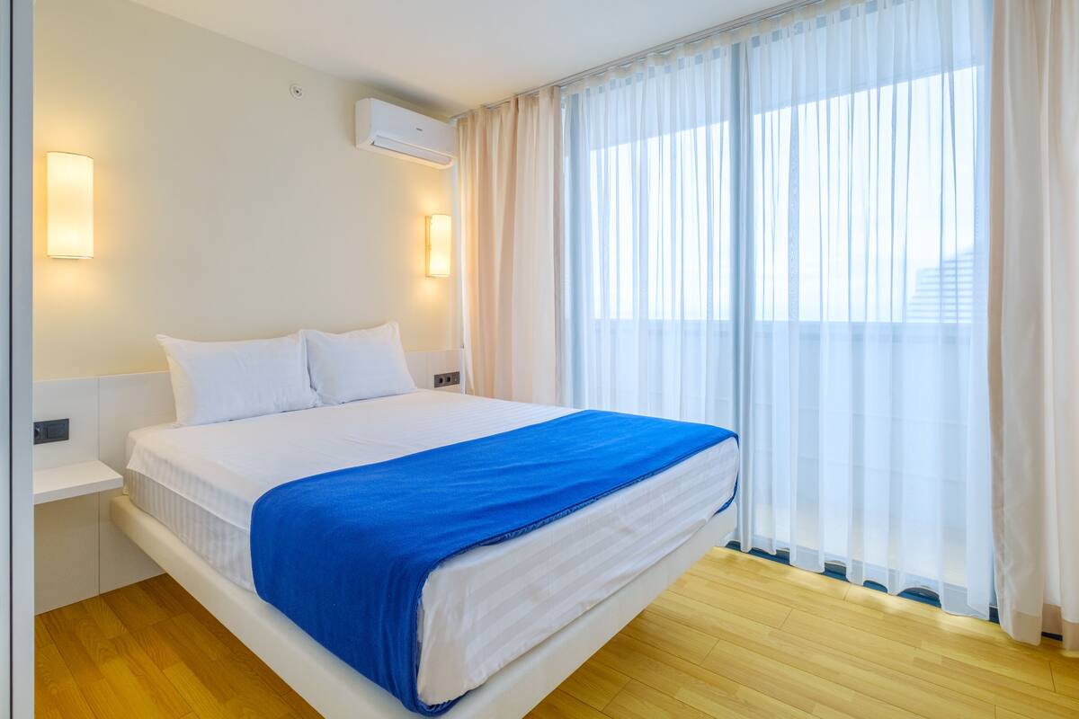 Una habitación de hotel con cama, paredes blancas, una ventana con cortinas de gasa y suelo de madera.