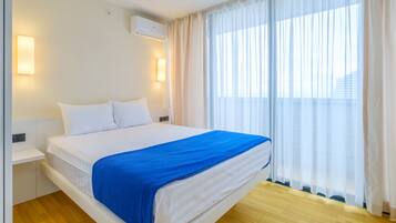 Una habitación de hotel con cama, paredes blancas, una ventana con cortinas de gasa y suelo de madera.