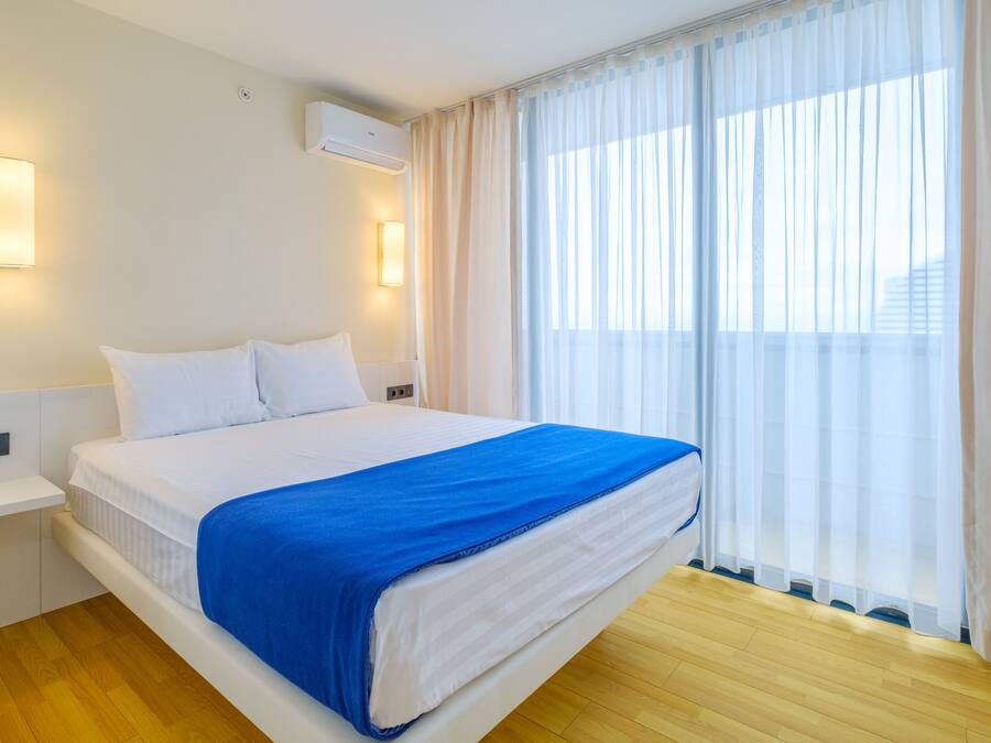 Una habitación de hotel con cama, paredes blancas, una ventana con cortinas de gasa y suelo de madera.