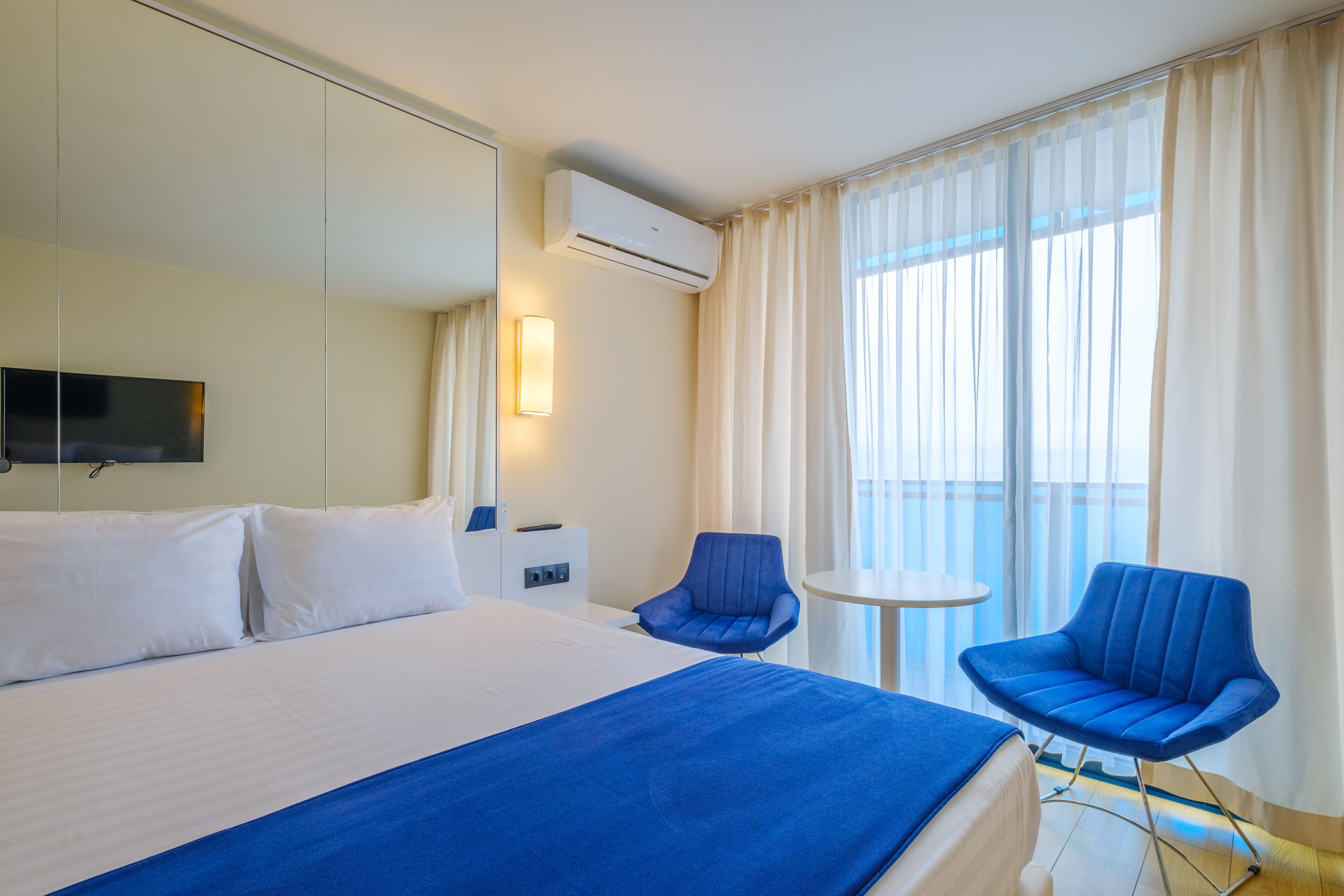 Habitación de hotel con una cama grande, dos sillas azules, una mesita, un televisor y un aire acondicionado.