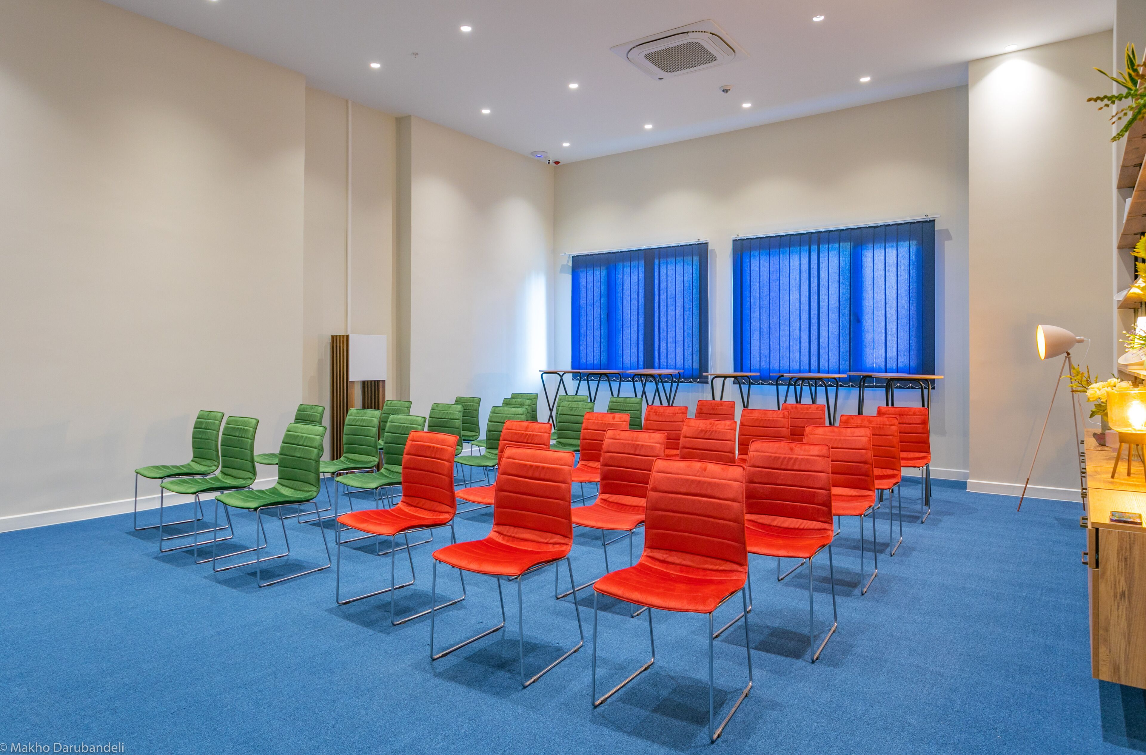 Un salón de conferencias con hileras de sillas verdes y rojas, alfombra azul y una ventana con cortinas azules.