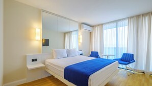 Una habitación de hotel moderna con una cama grande, una silla azul, una mesita pequeña y un espejo amplio.